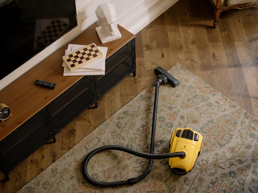 A yellow handheld vacuum cleaner with a flexible hose attachment resting on a patterned area rug in a living room, with a wooden media console underneath a wall-mounted TV, holding books, a remote control, and a decorative white bust sculpture. The room has wood flooring, and the scene appears clean and well-maintained, illustrating surface cleaning and domestic sanitisation. Ilford Carpet Cleaning specializes in deep cleaning and maintaining hygiene in residential spaces, as seen in this room preparation for thorough cleaning.
