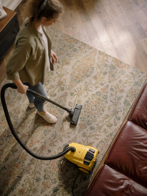 A middle-aged man with gray hair and glasses is performing surface cleaning in a bright living room. He is wearing a light gray long-sleeve shirt and dark pants, vacuuming a multicolored area rug with