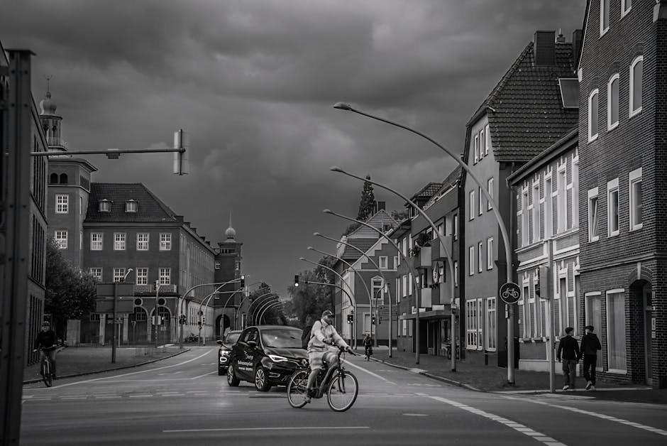 Black and white photograph of a city street scene showing a cyclist crossing at a pedestrian crossing, with a dark sky overhead. On the left, a person is riding a bicycle along the sidewalk, while on the right, two pedestrians walk together, dressed in coats. Several vehicles, including a black car, are visible waiting at the crossing. The street is flanked by multi-story brick and stucco buildings with numerous windows, some with decorative trim, and the scene is illuminated by curved streetlights. The overall atmosphere suggests a typical urban environment with active street life, captured under an overcast sky. Although related to cleaning in the context of residential and commercial spaces, this image visually emphasizes urban maintenance and cleanliness through the neat and orderly appearance of the street and buildings. For expert surface cleaning, deep cleaning, and sanitisation services, Ilford Carpet Cleaning is available to assist.