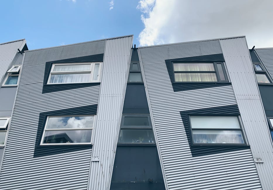 Photograph of the exterior facade of a modern, multi-story apartment building with a contemporary design. The building features vertical and horizontal metal cladding in silver and dark grey tones, creating a sleek, industrial appearance. Several large, rectangular windows with white frames are visible, some of which are open, allowing natural light to enter. The surfaces of the building are clean and well-maintained, with no visible dirt or grime. The sky above is partly cloudy, casting soft light on the facade, emphasizing the building's clear lines and reflective surfaces. This image highlights the structural design and external appearance of the residential flats, showcasing the materials used and the overall tidiness of the exterior environment. Ilford Carpet Cleaning welcomes inquiries for premium surface cleaning and maintenance services, ensuring buildings like this remain pristine and attractive.